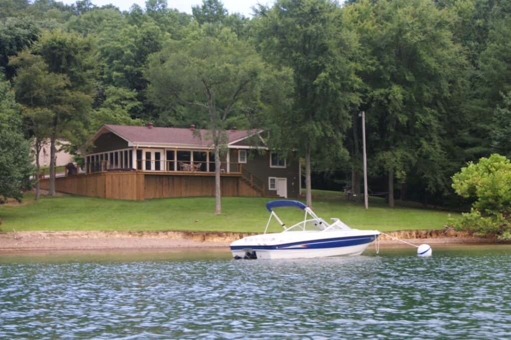 Ski boat attached to a buoy in front of a lake house nestled in green trees.
