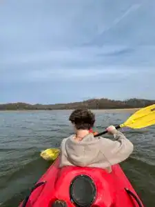 From the back of a red tandem kayak, a boy in the front seat is paddling out to the island.