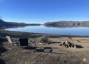 Carp fishing poles, chairs, and an unlit file line the shoreline of a glassy calm lake.