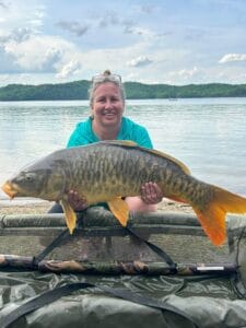 Woman smiling as she holds a large mirror carp on the shoreline with the lake in the background