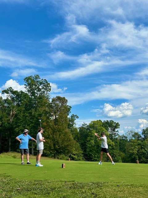 Three golfers, one driving, and two watching. Blue sky with clouds and green grass and trees.